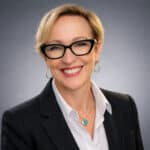 Headshot of Dr. Ac. Tammy Lalonde, CEO, wearing a black blazer, white collared shirt, and turquoise heart necklace against a soft gray studio background.