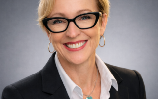 Headshot of Dr. Ac. Tammy Lalonde, CEO, wearing a black blazer, white collared shirt, and turquoise heart necklace against a soft gray studio background.