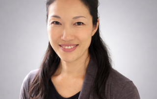 Headshot of Dr. Yoko Masuda, Acupuncturist, smiling softly in a dark top against a neutral gray background.