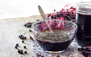 Elderberry syrup in a glass bowl and jar, placed on a floral towel atop a wooden surface, surrounded by elderberry branches.