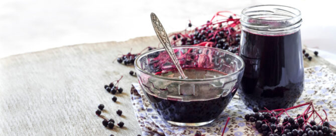 Elderberry syrup in a glass bowl and jar, placed on a floral towel atop a wooden surface, surrounded by elderberry branches.