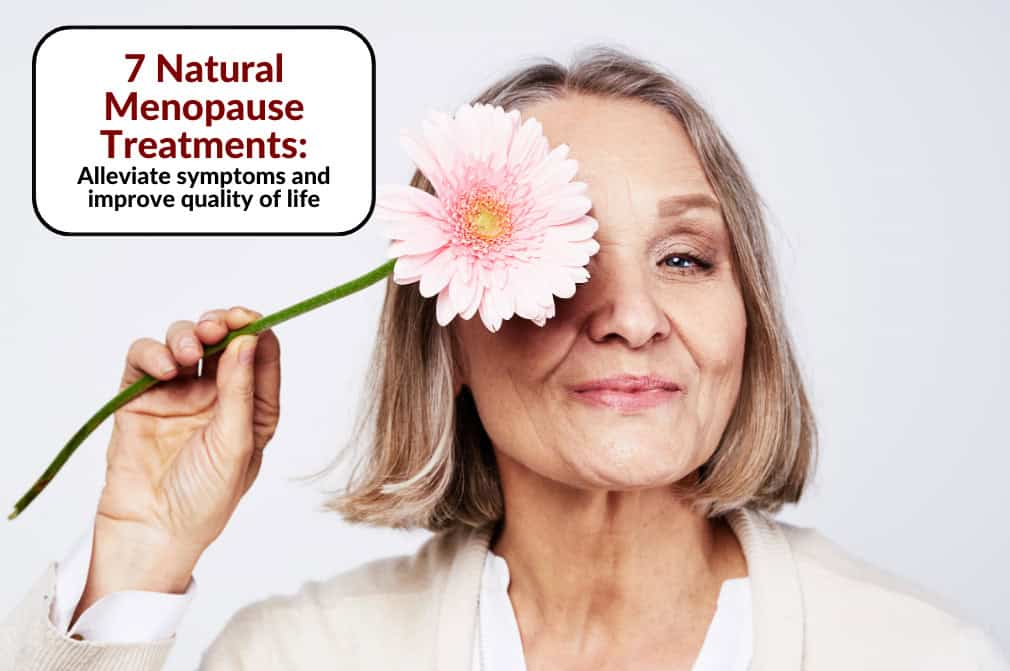 A smiling older woman holding a pink flower over one eye - Red Leaf Wellness Edmonton
