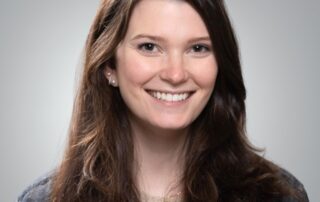 Portrait of Allie Kusnierczyk, smiling with long dark hair, wearing a denim jacket over a light-colored sweater against a neutral gray background.