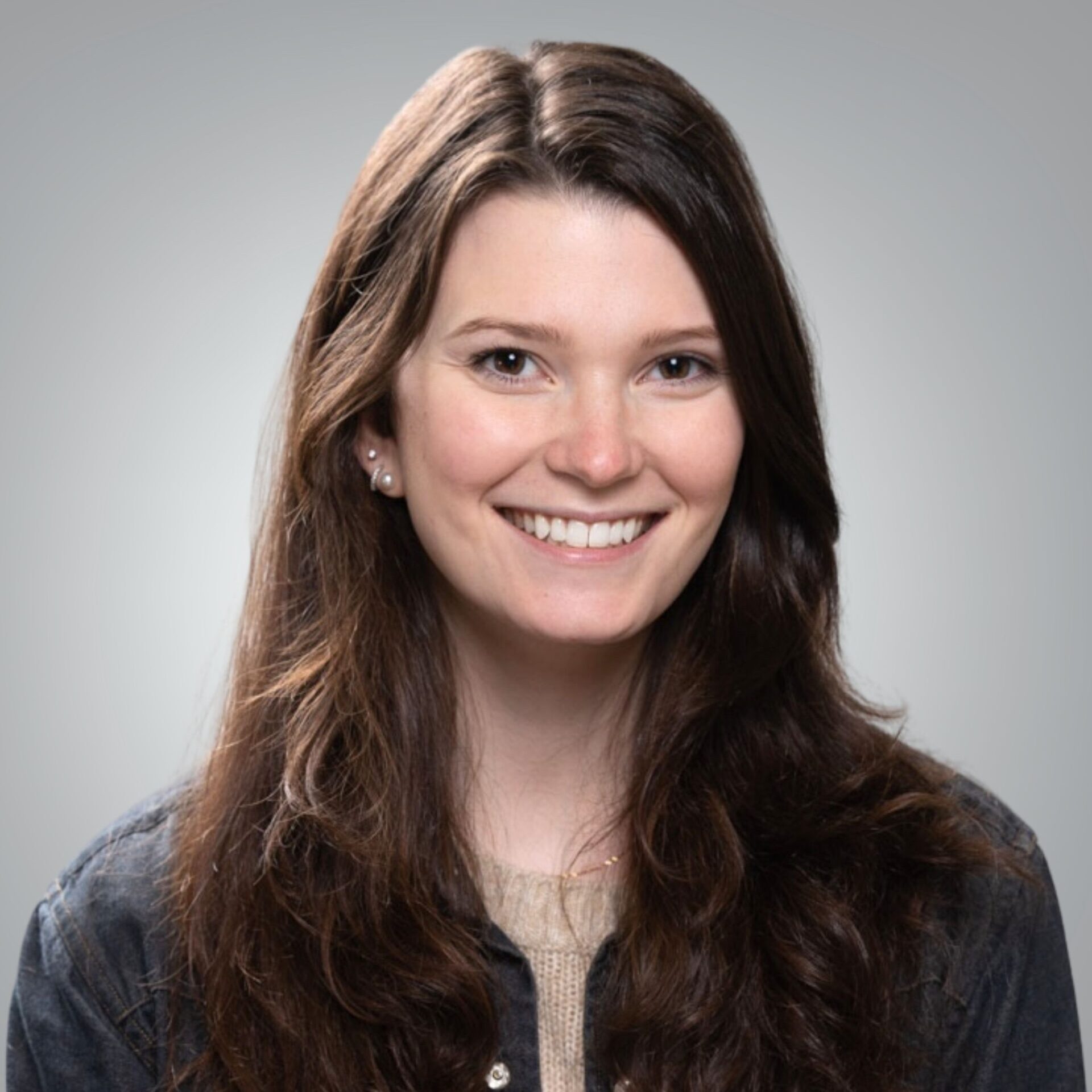 Portrait of Allie Kusnierczyk, smiling with long dark hair, wearing a denim jacket over a light-colored sweater against a neutral gray background.