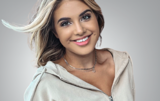 A friendly young woman with shoulder-length blonde hair, smiling warmly. She is wearing a light zip-up hoodie and a delicate necklace, exuding approachability and professionalism. The background is a soft gray gradient.