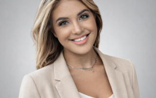 A friendly young woman with shoulder-length blonde hair, smiling warmly. She is wearing a light jacket and a delicate necklace, exuding approachability and professionalism. The background is a soft gray gradient.