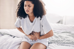 Young woman sitting on a bed and holding her stomach in pain, symbolizing health symptoms that could benefit from naturopathic support.