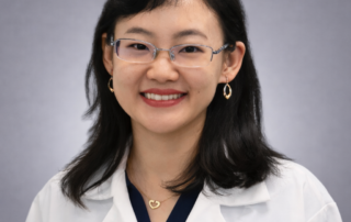 Headshot of Dr. Dan Jin, Acupuncturist, wearing glasses and a white clinical coat over a navy top against a soft gray studio background.