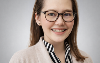 Professional headshot of Ulysssa (Uli) Urgel, wearing glasses and a white medical coat over a striped blouse, smiling against a soft gray background.