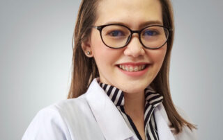 Professional headshot of Ulysssa (Uli) Urgel, wearing glasses and a white medical coat over a striped blouse, smiling against a soft gray background.