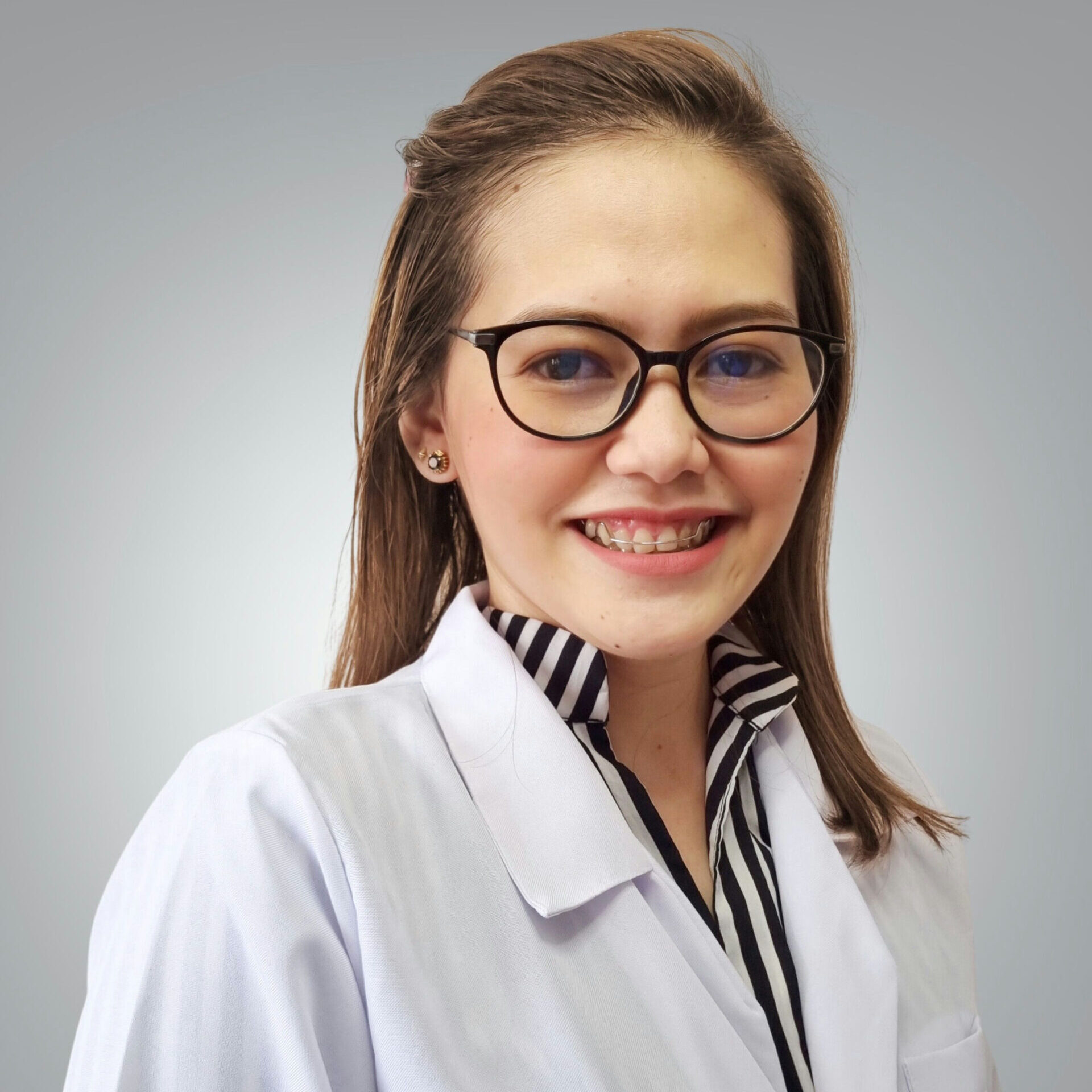 Professional headshot of Ulysssa (Uli) Urgel, wearing glasses and a white medical coat over a striped blouse, smiling against a soft gray background.