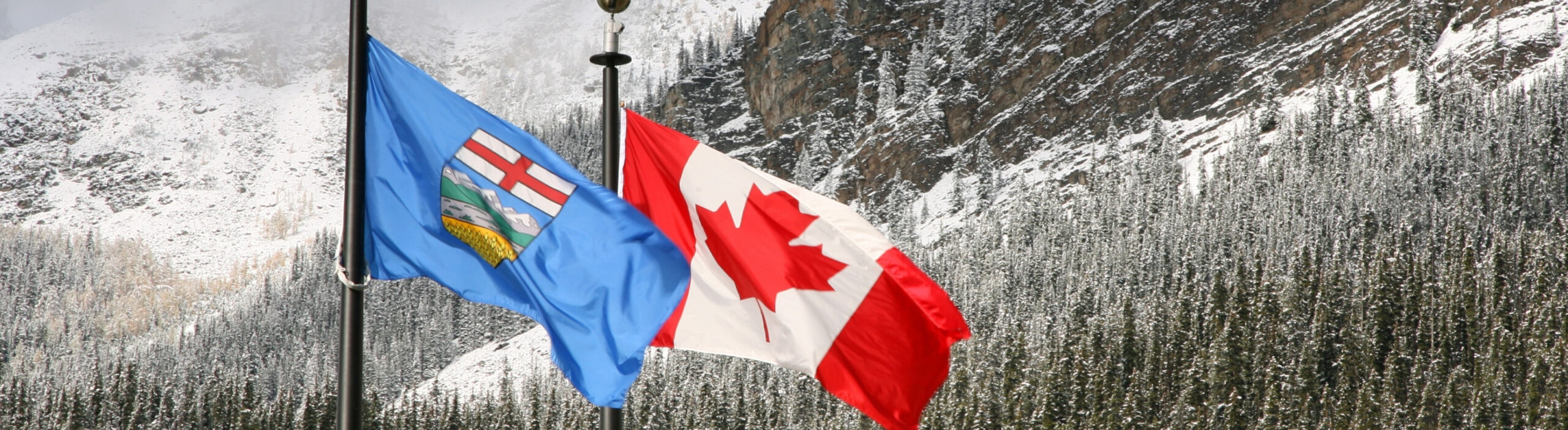 The Alberta and Canada flags flying side by side in front of a snow-dusted, forested mountain slope in the Canadian Rockies.
